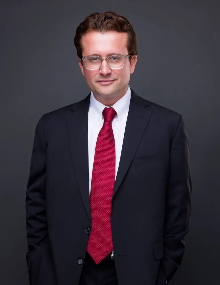 Luke Radney wearing glasses, a black suit with a white undershirt and a red tie.